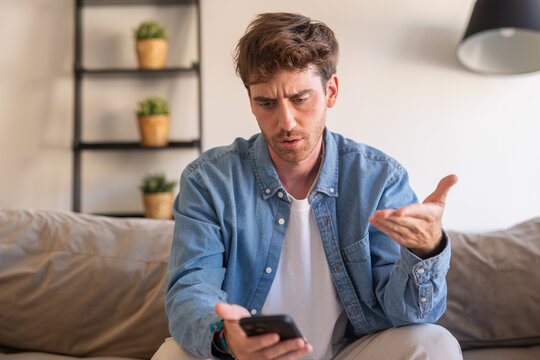 Young man on a sofa reacts with confusion and frustration to bad news on his smartphone, making a questioning gesture while scrolling through social media and messages at home
