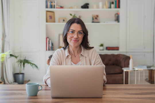 Young woman wearing eyeglasses sitting at a wooden desk with a laptop and coffee mug, smiling at the camera, embodying concepts of remote work, home office, and freelance lifestyle