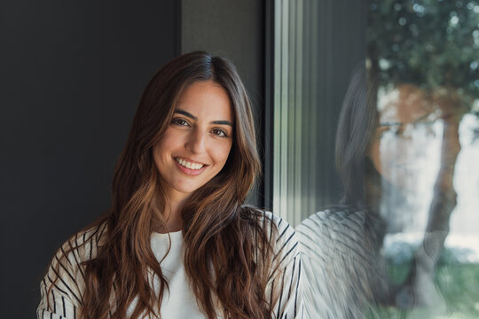 Happy young brunette woman smiling at camera by a large window with soft daylight and reflections, casual striped shirt, relaxed half-body portrait radiating warmth and confidence