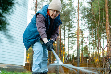 Fototapeta premium Senior woman raking fallen leaves in snowy backyard on cold winter day. Elderly homeowner clearing yard with long garden rake outdoors. Active retiree maintaining property despite winter weather