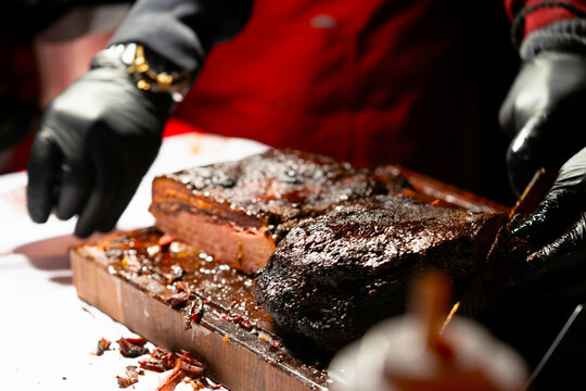 A juicy barbecued brisket being sliced on a wooden cutting board with a knife and tongs.
