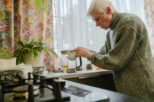 An elderly man with a noticeable stoop pours tea at kitchen counter. The scene illustrates persistence of daily habits despite physical limitations. Grandpa managing his drink.