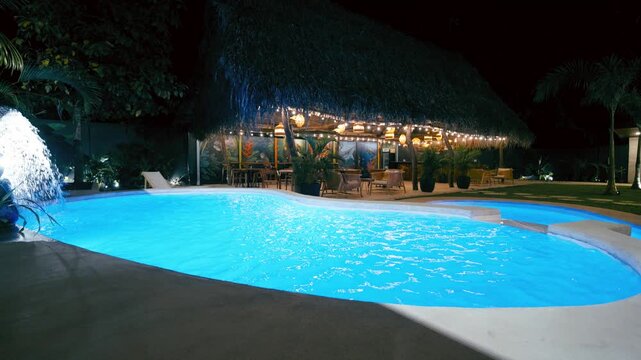 Luxurious swimming pool with glowing blue water at night. A serene hotel resort with a tropical tiki bar, waterfall, and lush garden