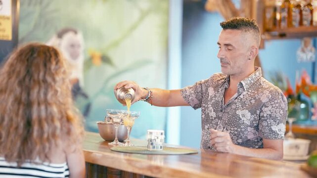 Experienced mixologist shaking and pouring a fresh tropical beverage into margarita glasses for a female customer at a beautiful exotic bar