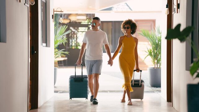 Stylish couple holding hands and walking with their suitcases down a modern hotel hallway, arriving for their vacation