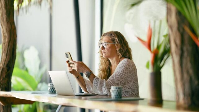 Digital nomad businesswoman with glasses working online on her laptop and scrolling on her smartphone in a beautiful tropical setting