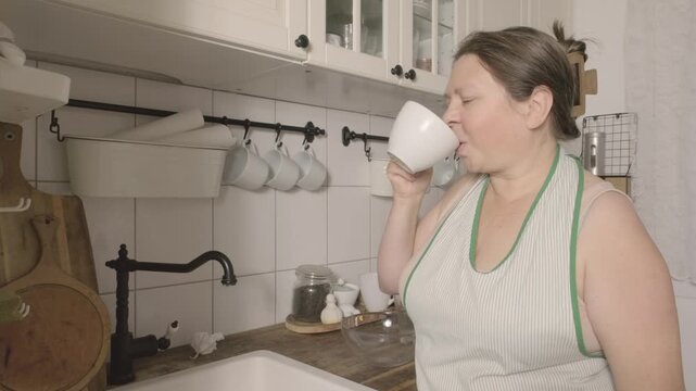 Woman in apron drinking water and resting in kitchen after cleaning, taking a break from housework, tired but satisfied, domestic routine, hydration, self care and everyday home life