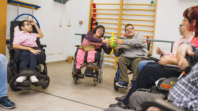 Group of adults with disabilities in wheelchairs engaging in rehabilitation exercises, passing a soft ball during physical therapy