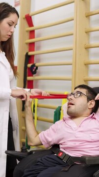Young man with a disability in a wheelchair performing physical therapy exercises. A female therapist is assisting him with a resistance band