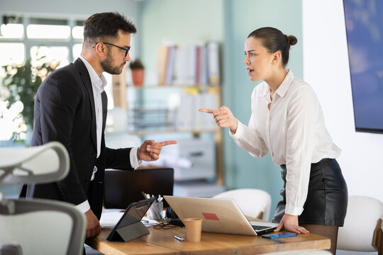 Young male and female standing on opposite sides of table and shouting at each other. Tense working atmosphere in office, employees resent and blame each other, quarrel or dispute.