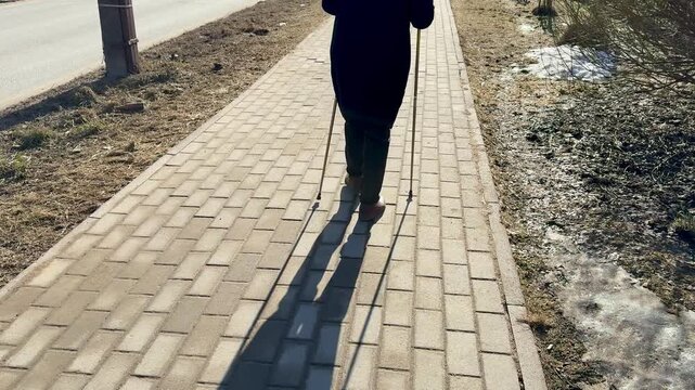 Woman Nordic walking shaded park path, brisk stride with poles and long shadow cast on paving, lea strewn verge, cool crisp light