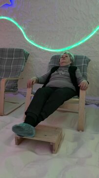 Elderly woman reclining in salt chamber, wearing cap and seated on wooden chair with footstool, legs extended over salt crystal