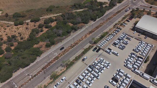 Aerial view of a car dealership with hundreds of new vehicles stored on rooftop lots alongside a highway in Israel, arid landscape background
