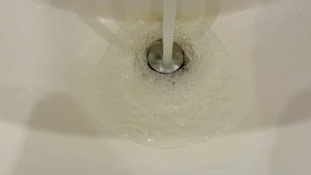 Dirty acrylic sink drain running water closeup showing yellowing stains, soap bubbles and scummy film swirling toward drain, foam