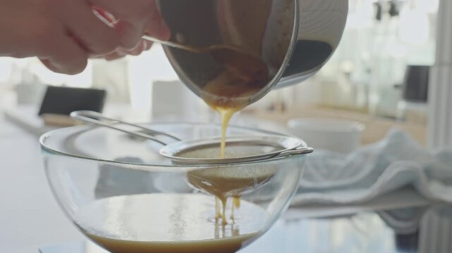 Person pours mixture from saucepan through sieve into glass bowl