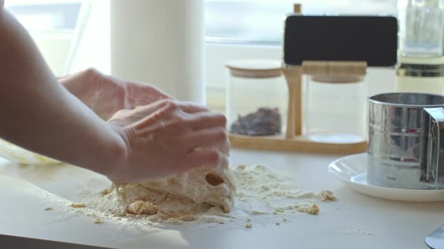 Hands knead dough with flour on kitchen counter near sifter and smartphone