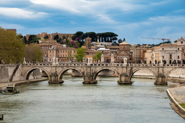 Fototapeta premium View on stone-arched bridge on Tiber river in Rome, balustrade, marble statues, cloudy sky, ochre and terracotta buildings, domes and umbrella pines rise on gently sloping Vatican hill