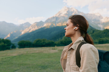 Fototapeta premium Woman outdoors, side profile looking toward mountains, backpack and light jacket, listening to music, sunlit meadow, natural colors, calm adventurous moment