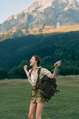 Fototapeta premium One person, a young woman hikes with a backpack through a vast green meadow, celebrating outdoors with towering mountains rising in the distance and a clear blue sky above.