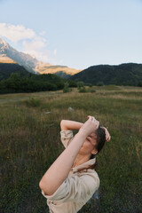 Fototapeta premium Young woman in a light jacket stands in a grassy field with distant mountains, arms raised, enjoying fresh air and warm sunlight in nature.