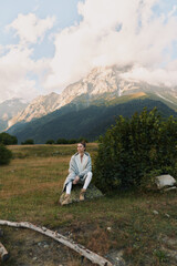 Fototapeta premium Young woman in casual clothes sitting on a rock in a mountain meadow with cloudy sky. Concept of lifestyle, nature, and outdoor relaxation in fresh air.