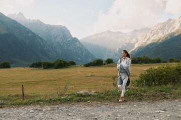Fototapeta premium A woman walks along a gravel path in a mountain valley, wearing a light shawl, enjoying outdoor lifestyle amid serene nature and expansive scenery, capturing a calm travel moment