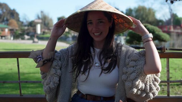 Cheerful young woman sitting on a bench in a japanese garden, smiling and playing with a conical hat