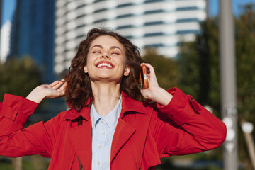Smiling woman in a red coat outdoors, phone to her ear, sunlit urban background, casual street...