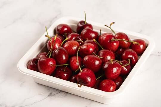 group of ripe, glossy red cherries packed in White Biodegradable paper tray on marble surface.  grocery marketing, healthy snack advertising, and fresh produce packaging .