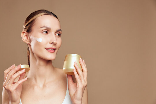 Young woman applying face cream with gentle smile, natural skin care routine, soft lighting, neutral background, healthy glowing complexion, beauty concept