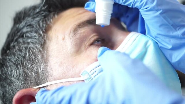 A medical professional in blue gloves carefully administers liquid drops into the eye of a male patient wearing a surgical mask.