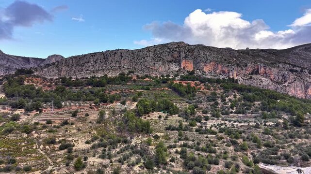 Aerial red cliff with terraced foothills and scrubby vegetation, expansive rocky escarpment against partly
