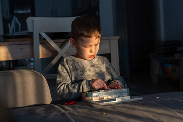 Fototapeta premium Little boy sorting colorful small pieces in plastic organizer box at table in dramatic golden light at home. Concept of fine motor skills development, early childhood learning and patience.