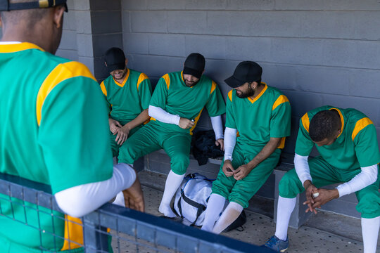 Diverse male baseball players sitting on dugout bench, wearing green uniforms, holding duffel bag