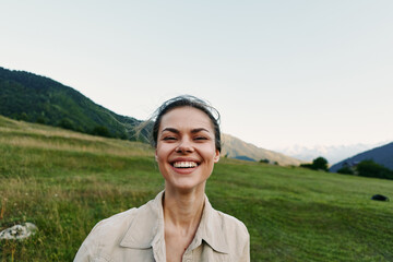 Fototapeta premium Bright smiling young woman stands in a green countryside field, wearing a light beige jacket, enjoying warm sun and open space, a candid moment of everyday wellbeing