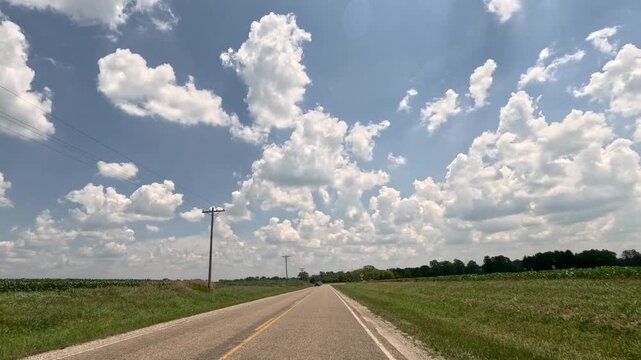 Slow-motion driving POV on a rural two-lane Midwest highway, passing oncoming vehicles towing a recreational boat and jet ski. Features open fields and a dramatic summer sky.