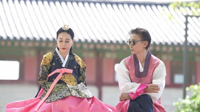 Two women in traditional Korean attire sit side by side. One wears a patterned top with a vibrant pink skirt. The other dons a simpler outfit with matching pink accents