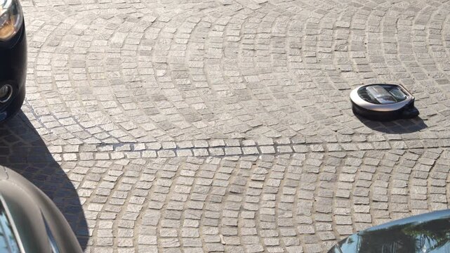 Modern robot vacuum cleaner placed drives along on gray cobblestone pavement outdoors, smart home gadget concept, lost technology scene, urban street setting, uprising of the machines