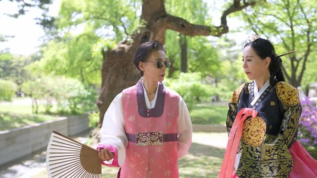 Two women in traditional Korean attire stroll through a park. One holds a folding fan, the other carries a pink sash. They chat, smiling, enjoying the bright, leafy surroundings