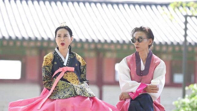 Two women in traditional Korean attire sit side by side. They wear vibrant, patterned robes with pink sashes. One woman speaks, the other listens, eyes closed. Their expressions show calm engagement