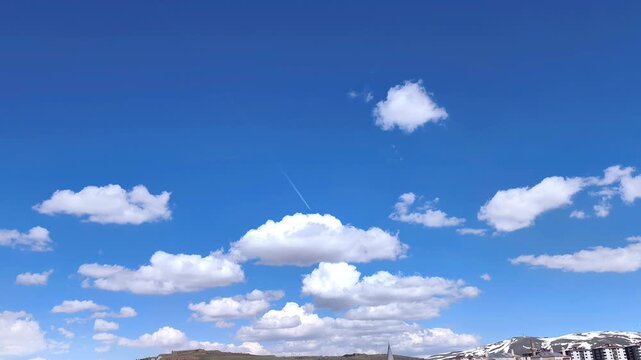 Static wide shot of a bright blue sky over Erzurum with fluffy white clouds and a long airplane contrail