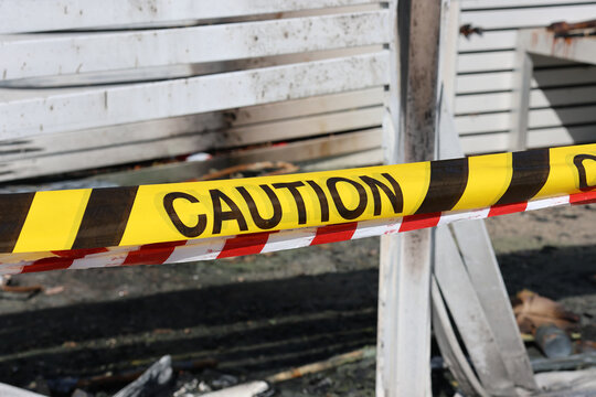 Yellow and black "Caution" tape cordoning off a damaged area following a fire, with charred structures and debris visible in the background