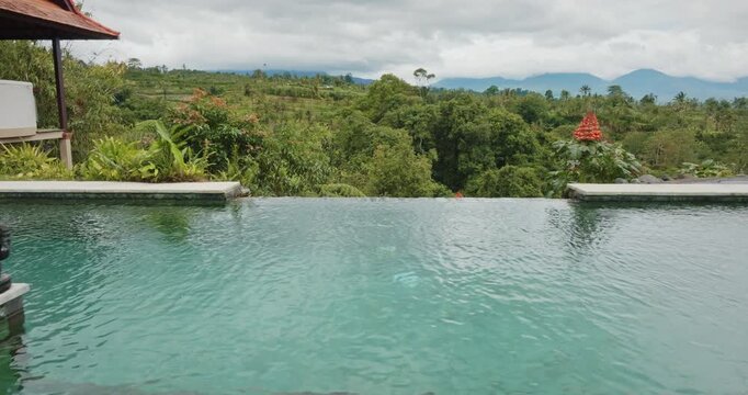 Bali infinity pool at luxury resort, serene views over lush tropical jungle, rice terraces, and distant mountains under cloudy sky, accented by traditional Balinese statues. Summer travel and relax