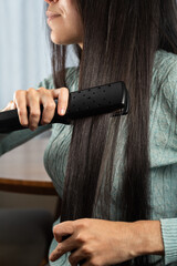 Naklejka premium Elegant woman straightening dark long hair with a styling tool during a relaxing beauty routine at home.
