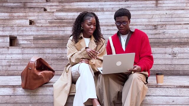 Cheerful african american couple friends man and woman sitting watching media on laptop outdoors in lounge area, smiling talkative black male and female buddies lively discussing video and joking.