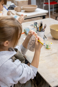Sketching pottery. Ceramic therapy. Talented kids girls learning creative hobby painting on earthenware cup using tool in artisan workshop.