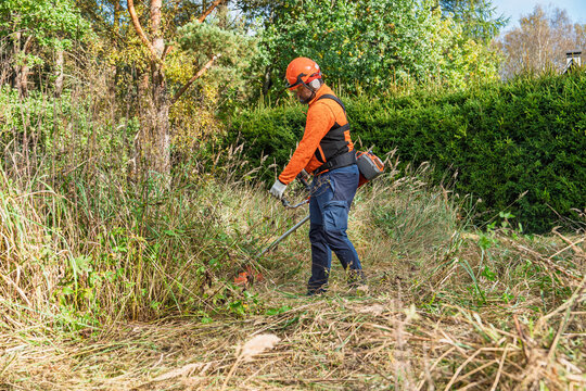 A man in Sweden is using a string trimmer to cut overgrown grass and brush in a yard. He is wearing safety gear, including a helmet, eye protection, and ear protection, for safe.