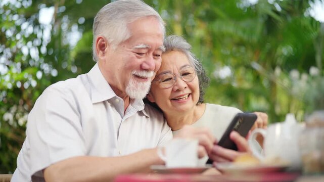 Happy Asian senior couple using smartphone together in garden at home, elderly husband and wife smiling while browsing mobile phone outdoors, active retirement lifestyle and joyful aging gracefully.