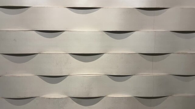 The curved wall panels form rhythmic, repeating waves. The smooth surface shimmers under soft, even lighting. Each curve casts soft, dark shadows. A backdrop for modern architecture or interior design