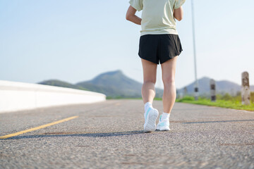 Naklejka premium Young adult Asian woman jogging and walking in the park at morning. Female athlete in sport shoes running on the road with visible leg muscles. Healthy lifestyle, wellness and workout concept